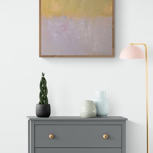 May include: A gray dresser with three drawers and gold knobs. The dresser is in front of a white wall with a framed abstract painting above it. There are two decorative vases and a potted plant on top of the dresser. A pink lamp with a gold base is on the right side of the image.