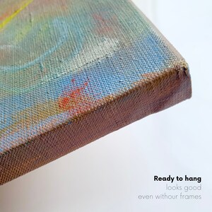 May include: Close-up of the edge of a canvas painting. The canvas is stretched over a wooden frame and the edges are painted a brown color. The painting is abstract with blue, green, yellow, and orange colors. The text "Ready to hang looks good even without frames" is visible at the bottom of the image.