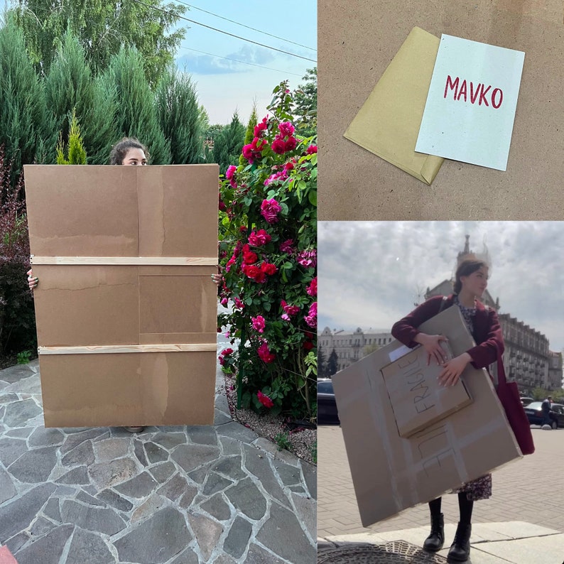 May include: A person holds a large cardboard box in front of a bush of pink roses. The box is brown and has three wooden strips across it. The person is wearing a white shirt and black pants. The box is being used to package a painting or artwork.