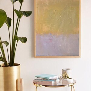 May include: A framed abstract painting with a gold and purple color scheme. The painting is hanging on a white wall above a gold side table with books and a small plant.