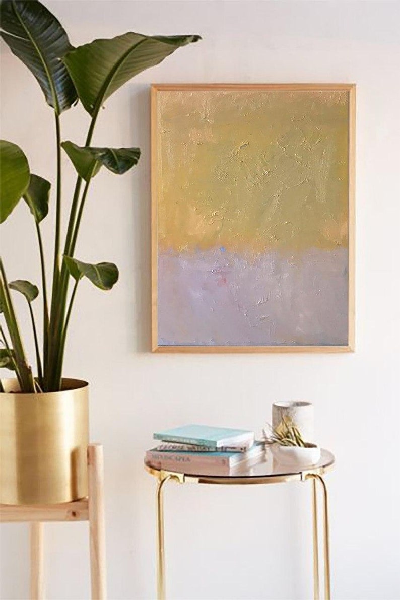May include: A framed abstract painting with a gold and purple color scheme. The painting is hanging on a white wall above a gold side table with books and a small plant.