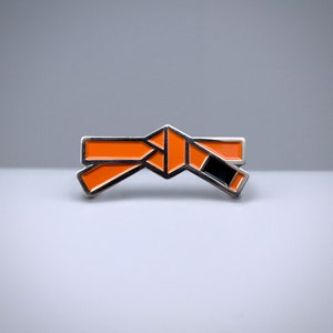 May include: Orange and black enamel pin shaped like a tied martial arts belt.