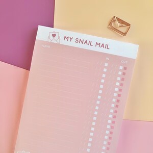 Può includere: Un blocco note rettangolare con sfondo rosa chiaro che riporta il testo MY SNAIL MAIL nella parte superiore. La pagina include colonne per nome, in e out con caselle di controllo. Il bordo inferiore mostra disegni a linee bianche di buste.