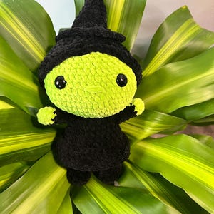 Pode incluir: Boneca de bruxa de crochê feita à mão com um rosto verde limão, olhos pretos e um chapéu e vestido de bruxa pretos. A boneca é colocada em um fundo de folhas verdes e amarelas, criando uma cena lúdica e festiva.