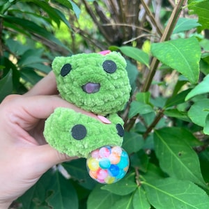 May include: A green crocheted plush toy featuring two stacked rounded segments with black circular eyes and a small purple mouth. A clear spherical charm filled with small, multicolored beads is attached to the bottom segment of the plush.