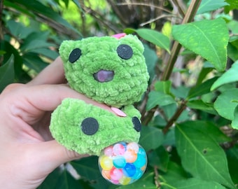 Pode incluir: Um brinquedo de pelúcia feito de crochê verde com dois segmentos arredondados empilhados, olhos circulares pretos e uma pequena boca roxa. Um pingente esférico transparente preenchido com pequenas contas multicoloridas está fixado na base.