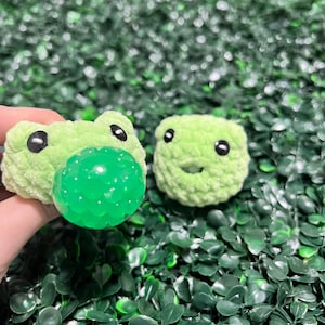 May include: Two green plush frog toys with black eyes. One frog has a green squishy ball attached to its mouth.