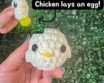 May include: A white crocheted chicken toy with black eyes and a yellow beak. The chicken is holding a yellow egg in its hand. The text "Chicken lays an egg!" is written above the chicken.
