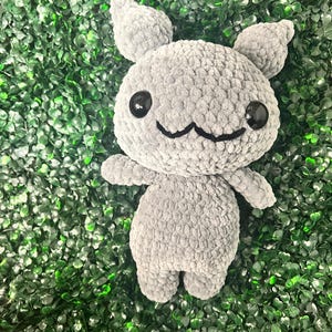 May include: A gray crocheted stuffed animal with large black eyes, small arms, and ears. The plush toy has a smiling face and is set against a green background.