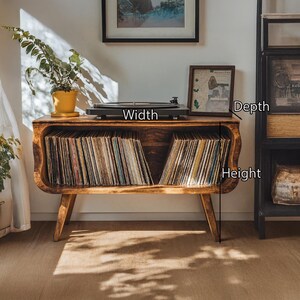 May include: A wooden record player stand with a built-in storage compartment for vinyl records. The stand is a warm brown color and has a curved design. A black record player sits on top. The stand is in a room with a plant and artwork.