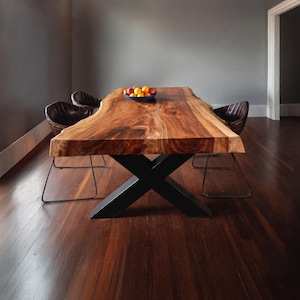 Könnte beinhalten: Ein großer Esstisch aus Holz mit natürlicher Kante und dunkelbraunem Finish. Der Tisch wird von einem schwarzen, X-förmigen Metallfuß getragen. Zwei Stühle mit braunen Ledersitzen sind zu sehen. Eine Schale mit Obst steht auf dem Tisch.