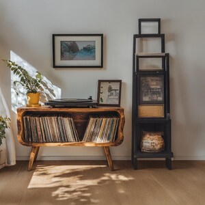 May include: A wooden record player console with a curved front, holding vinyl records. A black record player sits on top, with a framed picture and a potted plant. A black framed shelf unit is to the right.