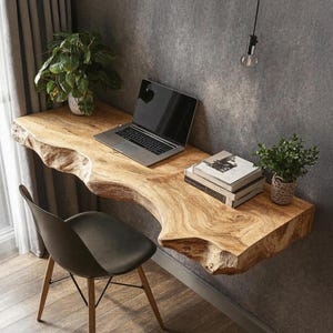 May include: A floating desk made from a single piece of live-edge wood, with a laptop, books, and a small potted plant. The desk is against a grey wall. A black chair with wooden legs is in front. A second plant in a white pot is on the desk.