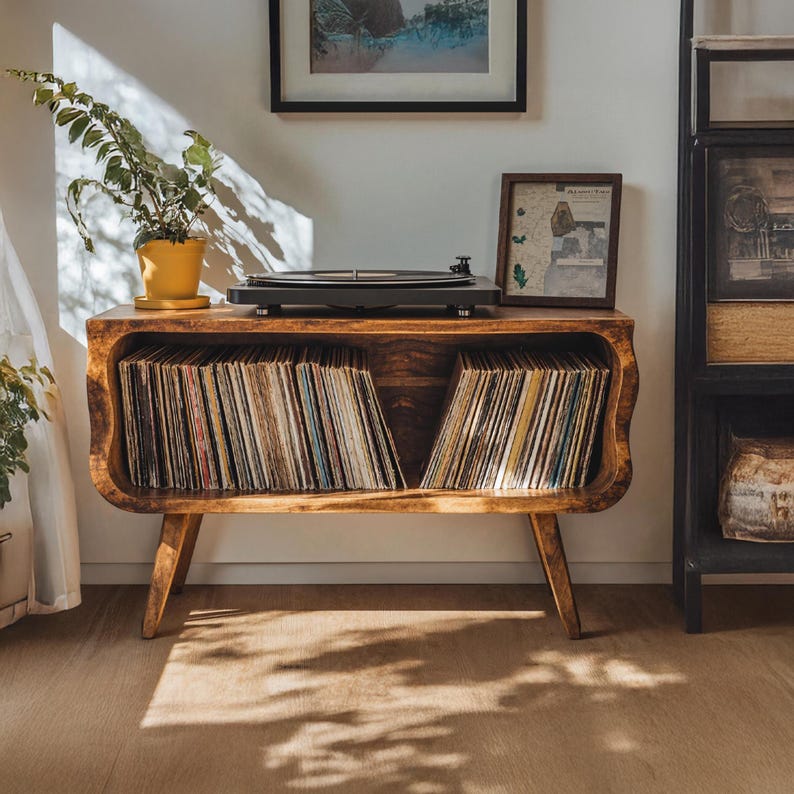 May include: A wooden record player console with a built-in storage compartment for vinyl records. The console has a mid-century modern design with angled legs and a curved front. A black turntable sits on top, and a potted plant is to the left.