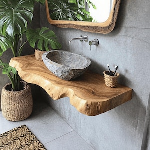 Puede incluir: Un tocador de baño con una encimera de madera natural y un lavabo de piedra. La encimera es una gruesa losa de madera con un borde vivo. El lavabo es un lavabo de piedra redondo y ovalado. El tocador está montado en la pared y tiene un espejo encima.