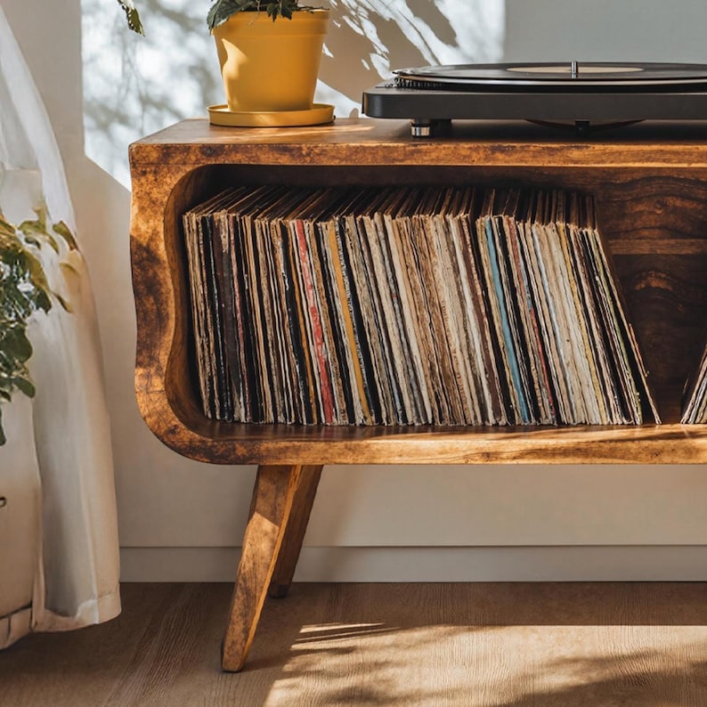 May include: A wooden record player stand with a curved design, holding a collection of vinyl records. A black record player sits on top, next to a yellow potted plant. The stand has angled legs and a warm, natural wood finish.