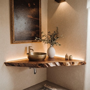 May include: A bathroom corner with a wooden countertop and a brass sink and tap. A white vase with greenery and small decorative bottles sit on the counter. A framed painting hangs above the sink.