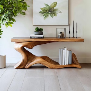 May include: A natural wood console table with a unique, organic design. The table features a flowing, sculptural base and a flat top. The table is holding books, a small potted plant, candles, and decorative items. The table is approximately 72 inches long.