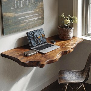 May include: A live edge wooden desk with a laptop, notebook, and a small plant in a woven basket. The desk is mounted on a wall and has a natural, organic shape. The laptop screen displays a tree branch image.