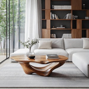May include: A modern living room featuring a light gray rug, a white sectional sofa, and a uniquely shaped wooden coffee table. The table has a natural wood grain finish and sits in front of a large window and a built-in wooden bookshelf.