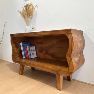 May include: A handcrafted wooden bookshelf with a unique, wavy design. The warm brown tones of the wood are complemented by the natural grain. Several books are visible inside the shelf. The bookshelf is supported by four short legs.