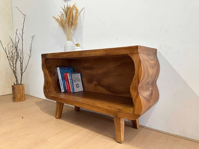May include: A handcrafted wooden bookshelf with a unique, wavy design. The warm brown tones of the wood are complemented by the natural grain. Several books are visible inside the shelf. The bookshelf is supported by four short legs.