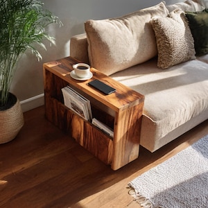 May include: A wooden side table with a magazine rack. The table is a warm brown colour and rectangular. A white coffee cup and a black smartphone sit on top. Magazines are visible in the rack. The table is likely made of solid wood.
