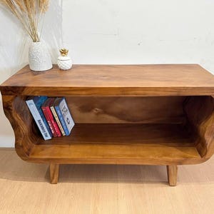 May include: A wooden, low-profile media console with a natural finish. The console features a shelf for storage and curved sides. Several books are placed inside the shelf. A decorative vase and pineapple-shaped container sit on top.