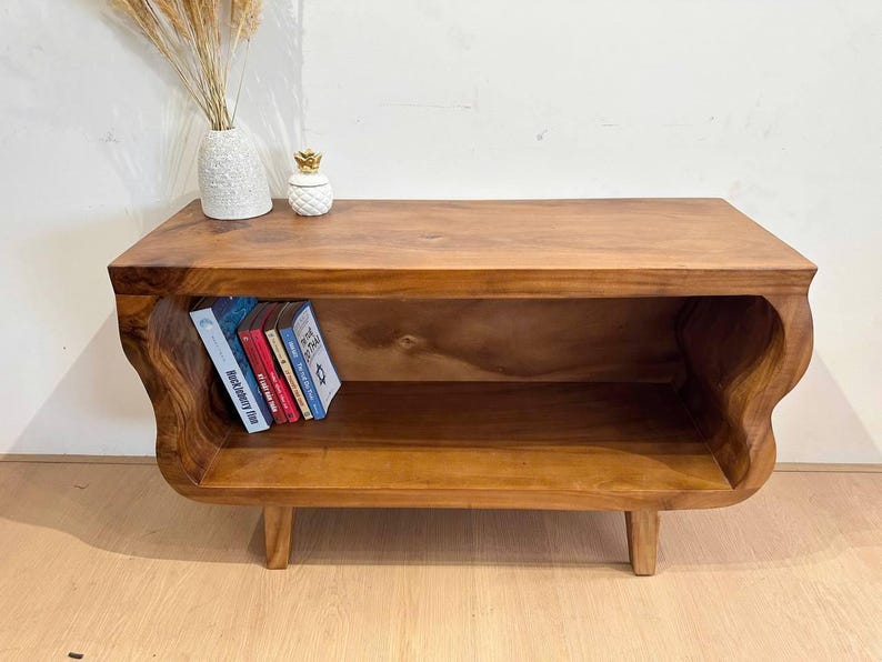 May include: A wooden, low-profile media console with a natural finish. The console features a shelf for storage and curved sides. Several books are placed inside the shelf. A decorative vase and pineapple-shaped container sit on top.