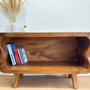 May include: A wooden bookshelf with a unique, wavy design. The warm brown tones of the wood are highlighted by the natural grain. Several books are placed inside the shelf. The shelf is supported by four short legs.