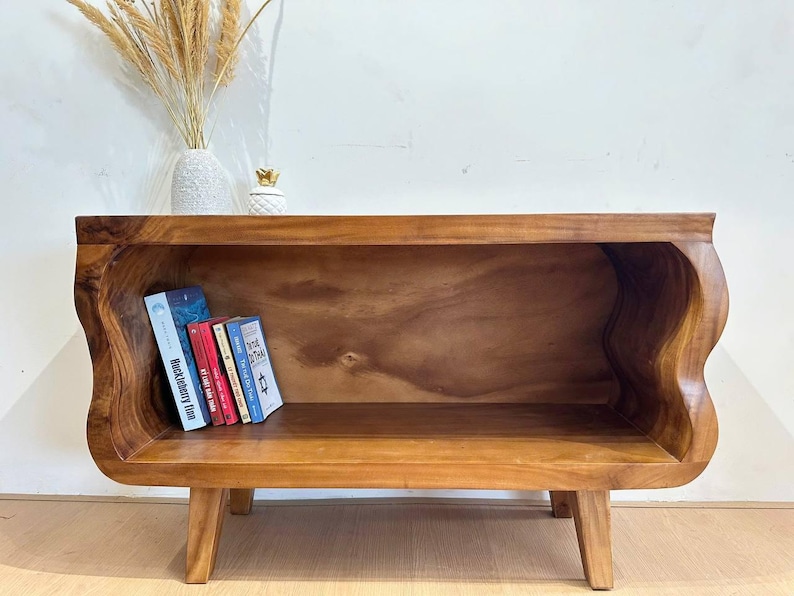 May include: A wooden bookshelf with a unique, wavy design. The warm brown tones of the wood are highlighted by the natural grain. Several books are placed inside the shelf. The shelf is supported by four short legs.