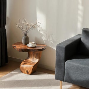 K&ouml;nnte beinhalten: Ein Holz-Beistelltisch mit einer einzigartigen, organischen Form. Der Tisch hat eine nat&uuml;rliche Holzoptik und ein geschwungenes, flie&szlig;endes Design. Auf dem Tisch stehen eine kleine wei&szlig;e Tasse und Untertasse sowie eine Vase mit Trockenblumen.