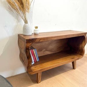 May include: A wooden, low-profile bookshelf with a natural finish. The shelf has a curved design on the sides and is supported by four short legs. Several books are visible inside the shelf. A vase with dried plants and a decorative pineapple are on top.