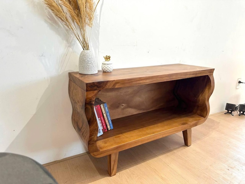 May include: A wooden, low-profile bookshelf with a natural finish. The shelf has a curved design on the sides and is supported by four short legs. Several books are visible inside the shelf. A vase with dried plants and a decorative pineapple are on top.