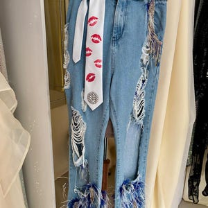 Pu&ograve; includere: Jeans di denim blu chiaro con dettagli strappati, una cintura bianca e una cravatta bianca stampata con labbra rosse. I jeans presentano finiture in piume blu alle caviglie e abbellimenti decorativi.