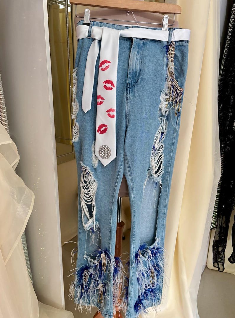 Pu&ograve; includere: Jeans di denim blu chiaro con dettagli strappati, una cintura bianca e una cravatta bianca stampata con labbra rosse. I jeans presentano finiture in piume blu alle caviglie e abbellimenti decorativi.