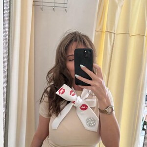 Pu&ograve; includere: Cravatta bianca con stampa di labbra rosse e un grande ornamento rotondo di strass scintillanti. La cravatta &egrave; annodata a fiocco. Viene indossata con una maglietta beige e jeans blu.