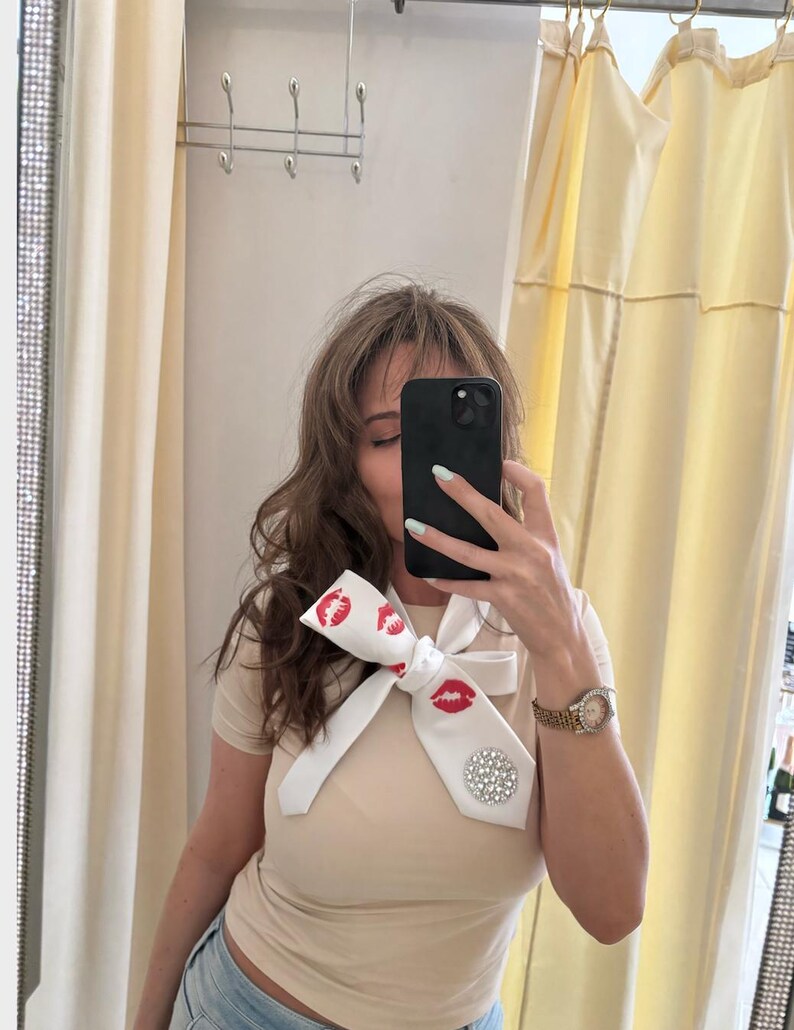 Pu&ograve; includere: Cravatta bianca con stampa di labbra rosse e un grande ornamento rotondo di strass scintillanti. La cravatta &egrave; annodata a fiocco. Viene indossata con una maglietta beige e jeans blu.