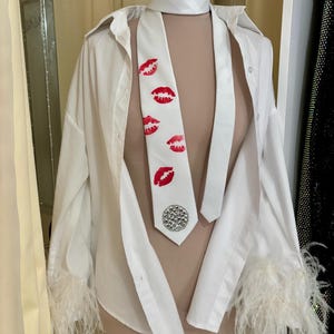 Pu&ograve; includere: Una camicia bianca con bottoni e finiture in piume sui polsini, abbinata a una cravatta bianca con stampe di labbra rosse e un abbellimento argentato. L'outfit &egrave; esposto su un manichino.