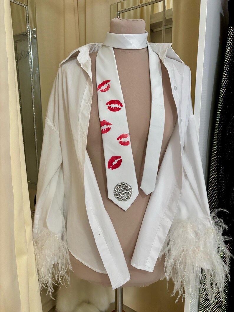 Pu&ograve; includere: Una camicia bianca con bottoni e finiture in piume sui polsini, abbinata a una cravatta bianca con stampe di labbra rosse e un abbellimento argentato. L'outfit &egrave; esposto su un manichino.