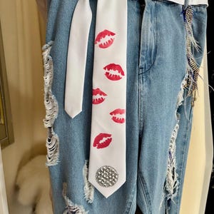 Pu&ograve; includere: Jeans di denim blu chiaro con dettagli strappati e una cravatta bianca con stampe di labbra rosse. La cravatta &egrave; adornata da un abbellimento di strass. &Egrave; visibile anche una cintura bianca.