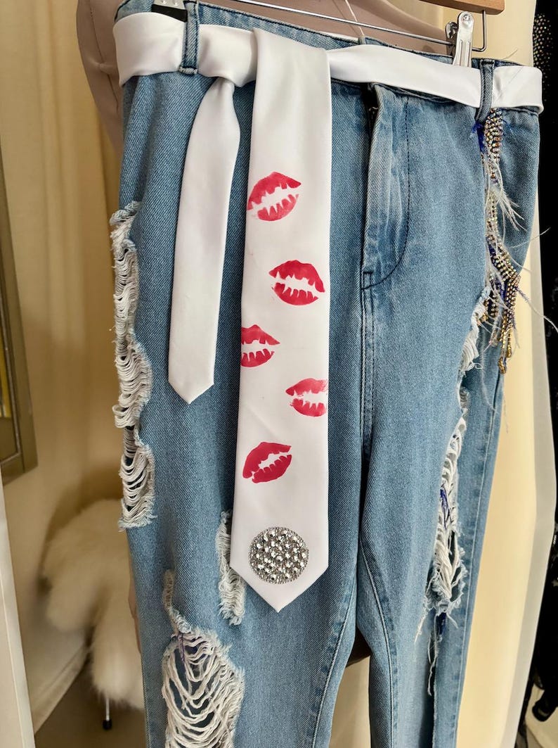 Pu&ograve; includere: Jeans di denim blu chiaro con dettagli strappati e una cravatta bianca con stampe di labbra rosse. La cravatta &egrave; adornata da un abbellimento di strass. &Egrave; visibile anche una cintura bianca.