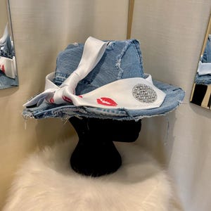 Pu&ograve; includere: Un cappello fedora in denim con tesa strappata. Il cappello &egrave; adornato con un nastro di raso bianco con stampe di labbra rosse e un abbellimento circolare di strass. Il cappello &egrave; esposto su una testa di manichino nera.