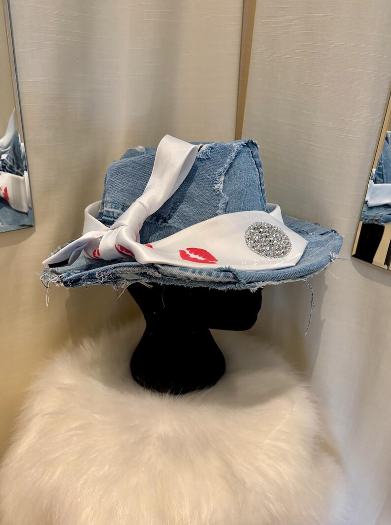 Pu&ograve; includere: Un cappello fedora in denim con tesa strappata. Il cappello &egrave; adornato con un nastro di raso bianco con stampe di labbra rosse e un abbellimento circolare di strass. Il cappello &egrave; esposto su una testa di manichino nera.