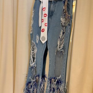 Pu&ograve; includere: Jeans di denim blu chiaro con dettagli strappati e cintura bianca. I jeans presentano sezioni strappate e sono adornati da piume blu sull'orlo. Una cravatta bianca con stampe di labbra rosse aggiunge un tocco unico.