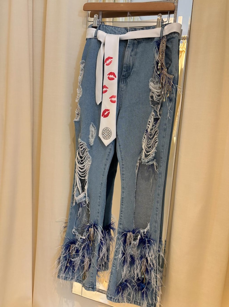 Pu&ograve; includere: Jeans di denim blu chiaro con dettagli strappati e cintura bianca. I jeans presentano sezioni strappate e sono adornati da piume blu sull'orlo. Una cravatta bianca con stampe di labbra rosse aggiunge un tocco unico.