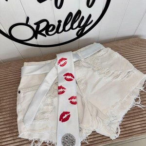 Pu&ograve; includere: Pantaloncini di jeans bianchi con dettagli strappati e cintura bianca abbinata. Una cravatta bianca con stampe di labbra rosse e un abbellimento di paillettes argentate &egrave; drappeggiata sui pantaloncini. &Egrave; visibile anche il testo "helifeof Reilly".