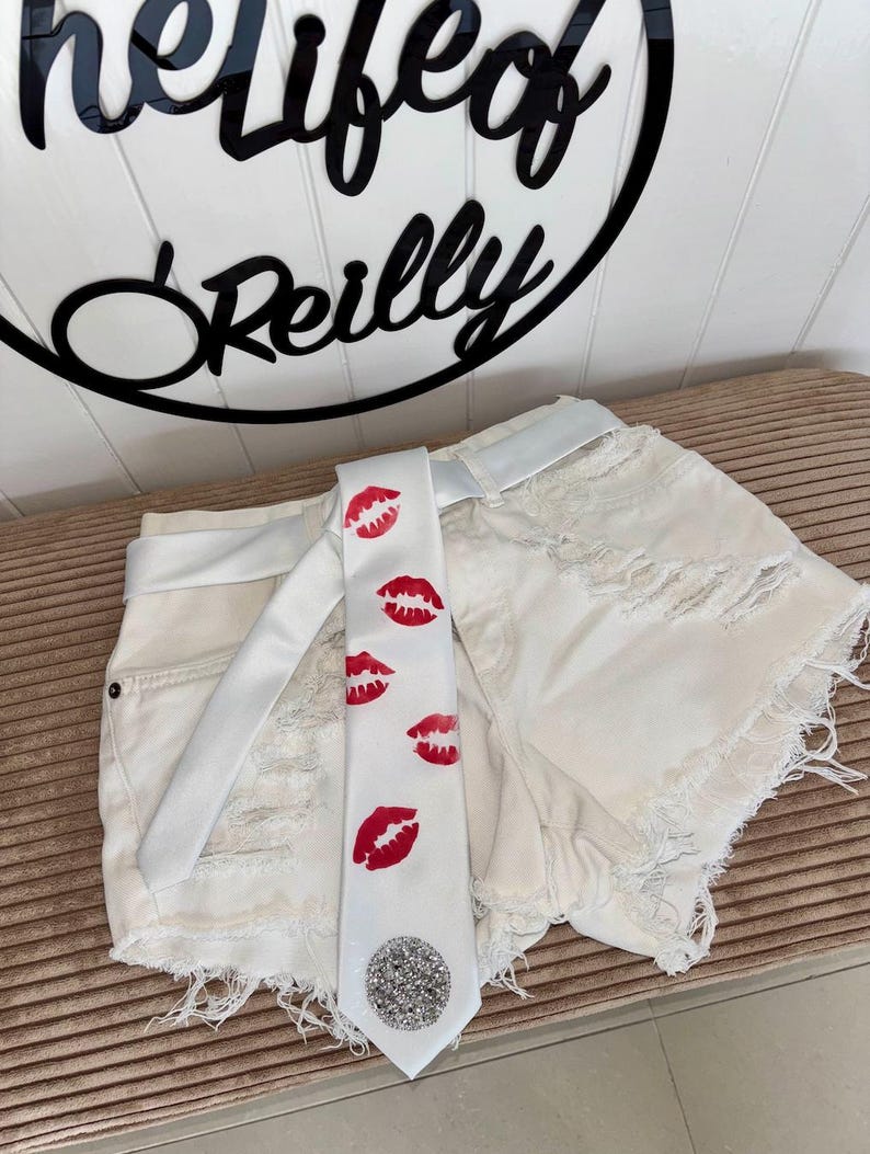 Pu&ograve; includere: Pantaloncini di jeans bianchi con dettagli strappati e cintura bianca abbinata. Una cravatta bianca con stampe di labbra rosse e un abbellimento di paillettes argentate &egrave; drappeggiata sui pantaloncini. &Egrave; visibile anche il testo "helifeof Reilly".