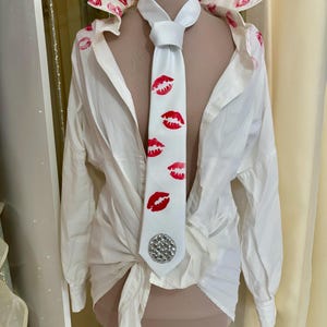 Pu&ograve; includere: Una camicia bianca con segni di rossetto rosso sul colletto e una cravatta bianca. La cravatta presenta stampe di baci di rossetto rosso e un abbellimento di strass argentati. La camicia &egrave; annodata in vita.