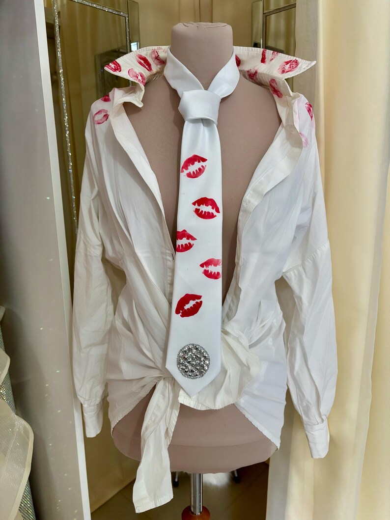 Pu&ograve; includere: Una camicia bianca con segni di rossetto rosso sul colletto e una cravatta bianca. La cravatta presenta stampe di baci di rossetto rosso e un abbellimento di strass argentati. La camicia &egrave; annodata in vita.